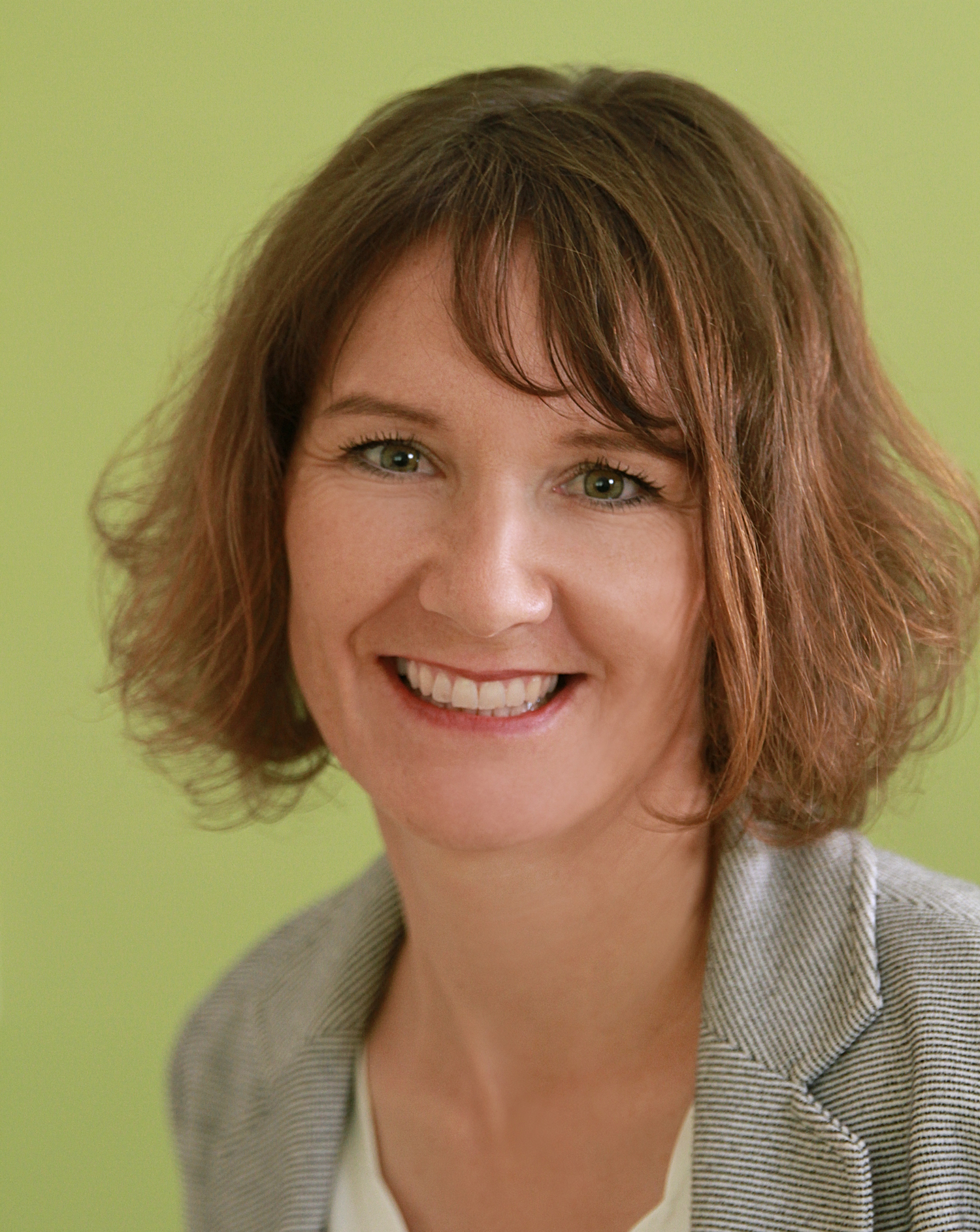 NIna Mattes, lächelt, trägt einen gestreiften Blazer und hat brünette mittellange Haare